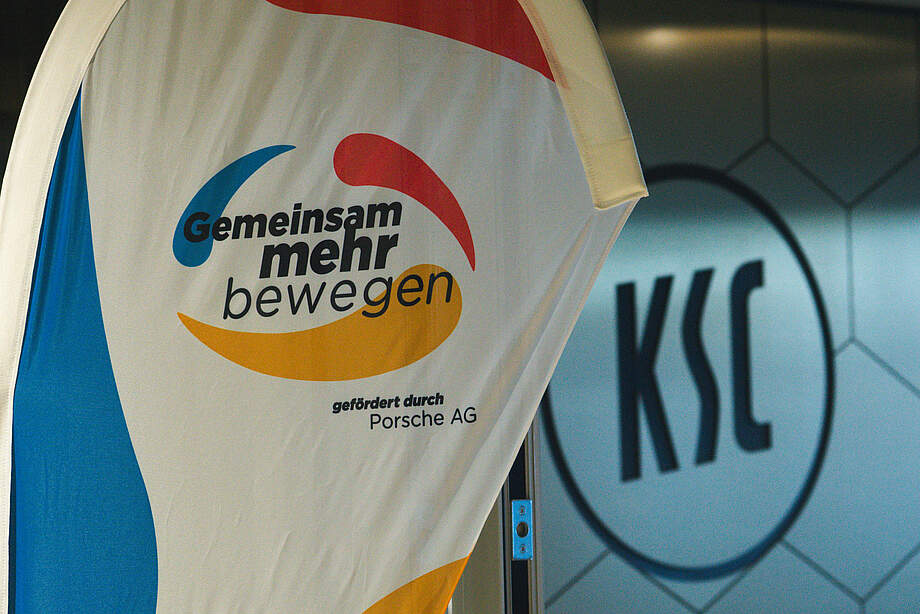 Banner mit dem Slogan 'Gemeinsam mehr bewegen' und dem Logo des Karlsruher SC im Hintergrund. Das Banner wird von der Porsche AG gefördert.