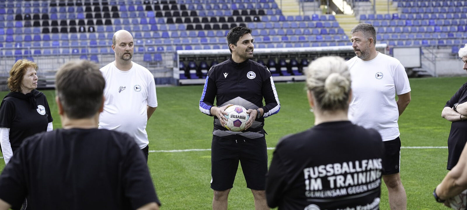 Eine Gruppe von Menschen steht auf einem Fußballfeld und hört einem Trainer zu, der einen Fußball in den Händen hält. Im Hintergrund sind leere Tribünen zu sehen. Eine Person trägt ein T-Shirt mit der Aufschrift "FUSSBALLFANS IM TRAINING".