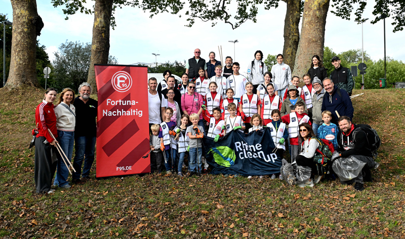 Eine große Gruppe von Menschen, darunter Spieler, Fans und Freiwillige, steht für ein Gruppenfoto auf einer Wiese. Sie tragen T-Shirts und Jacken in den Farben des Fortuna Düsseldorf-Logos und halten Besen und Müllsäcke in den Händen. Im Hintergrund sind Bäume und ein Fußballfeld zu sehen. Die Gruppe hält ein rotes Banner mit dem Text "Fortuna-Nachhaltig" und ein blaues Banner mit dem Text "Rhine Clean Up" hoch.