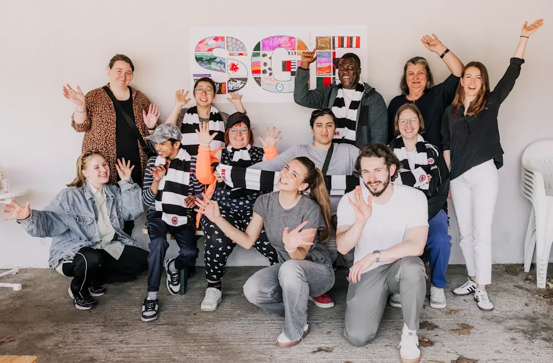 Eine Gruppe von Menschen, darunter Mitarbeiter und ein Spieler des Eintracht Frankfurt, posieren fröhlich für ein Gruppenfoto. Sie sitzen und stehen vor einem bunt gestalteten Hintergrund mit dem Logo der REWE Aktionstasse. Alle lächeln und winken in die Kamera, was eine positive und freundliche Atmosphäre vermittelt.