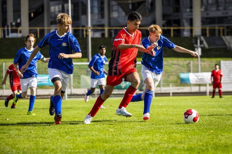 Jugendfußballspiel: Drei junge Fußballspieler in Aktion auf einem Rasenplatz. Ein Spieler im roten Trikot dribbelt den Ball, während zwei Spieler im blauen Trikot versuchen, den Ball zu erobern. Im Hintergrund sind weitere Spieler und eine Tribüne zu sehen.