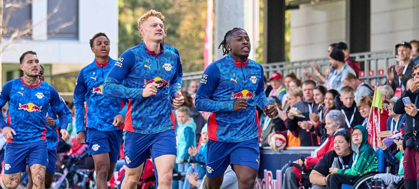 Spieler des RB Leipzig laufen auf den Platz und werden von jubelnden Fans begrüßt. Die Spieler tragen blaue Trikots mit roten und gelben Akzenten. Im Vordergrund ist eine Person im Rollstuhl zu sehen, die ebenfalls die Spieler begrüßt. Die Atmosphäre ist lebhaft und inklusiv.