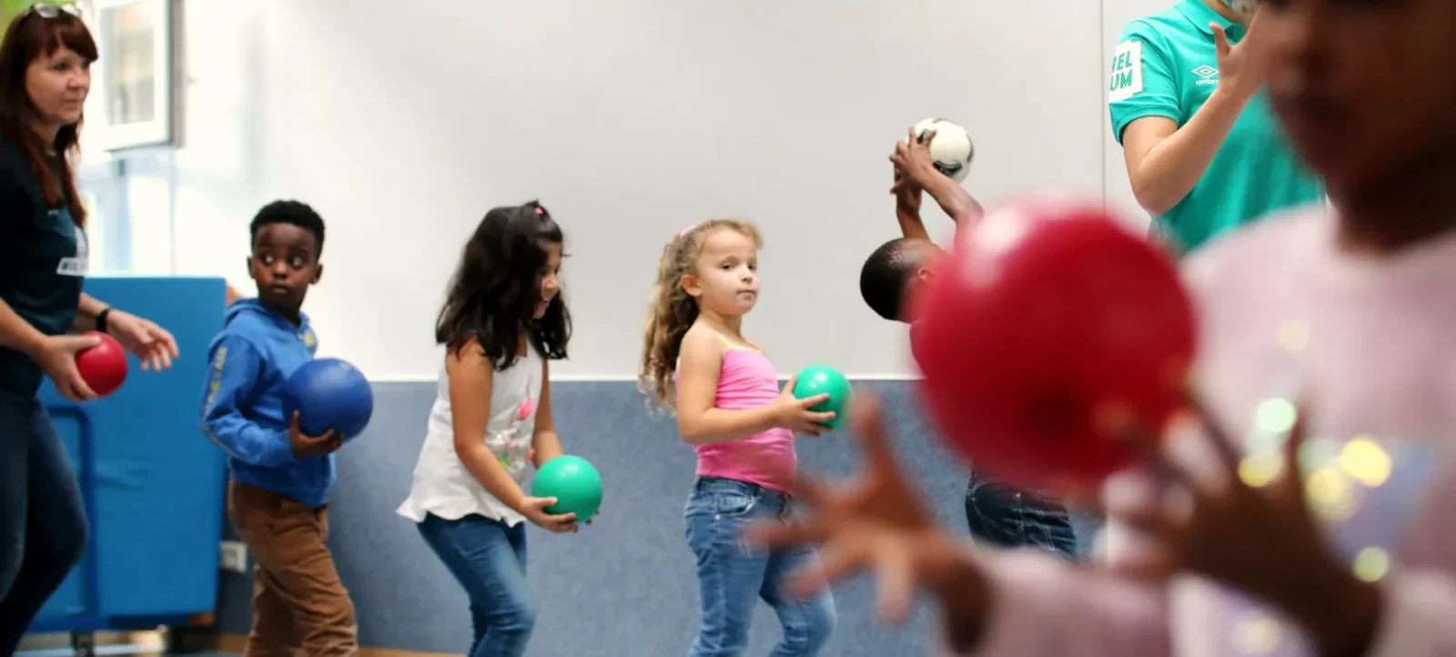 Kinder nehmen an einem Ballspiel in einer Sporthalle teil. Sie werden von zwei Erwachsenen angeleitet, die jeweils ein T-Shirt mit einem Vereinslogo tragen. Die Kinder halten bunte Bälle in den Händen und scheinen sich auf eine Übung mit einem Fußball zu konzentrieren.