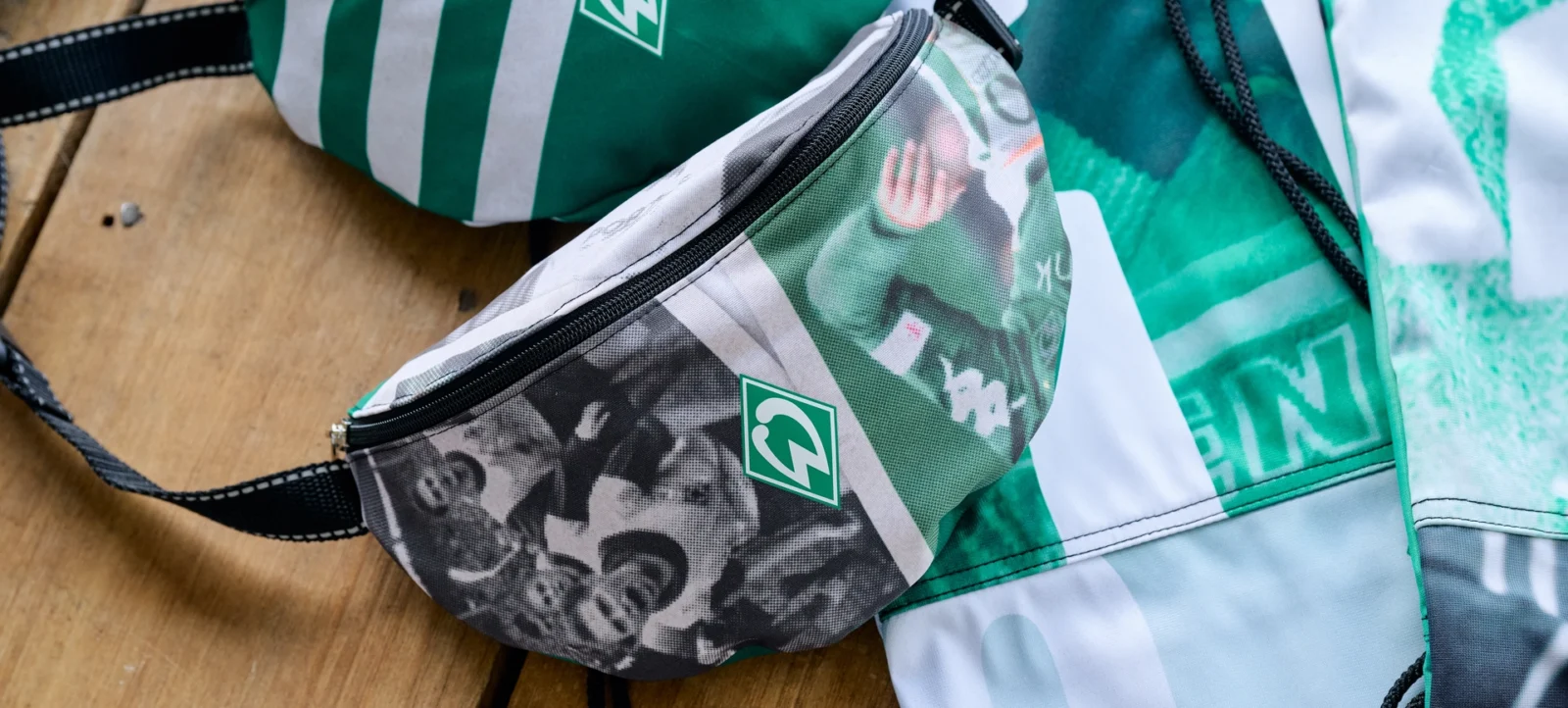 Ein Bild von zwei Stoffmasken und einer Tasche, die alle das Logo des Hertha BSC Vereins tragen. Die Maske im Vordergrund hat ein schwarz-weißes Muster, während die andere Maske und die Tasche grün-weiß gestreift sind. Alle Gegenstände liegen auf einem Holztisch.