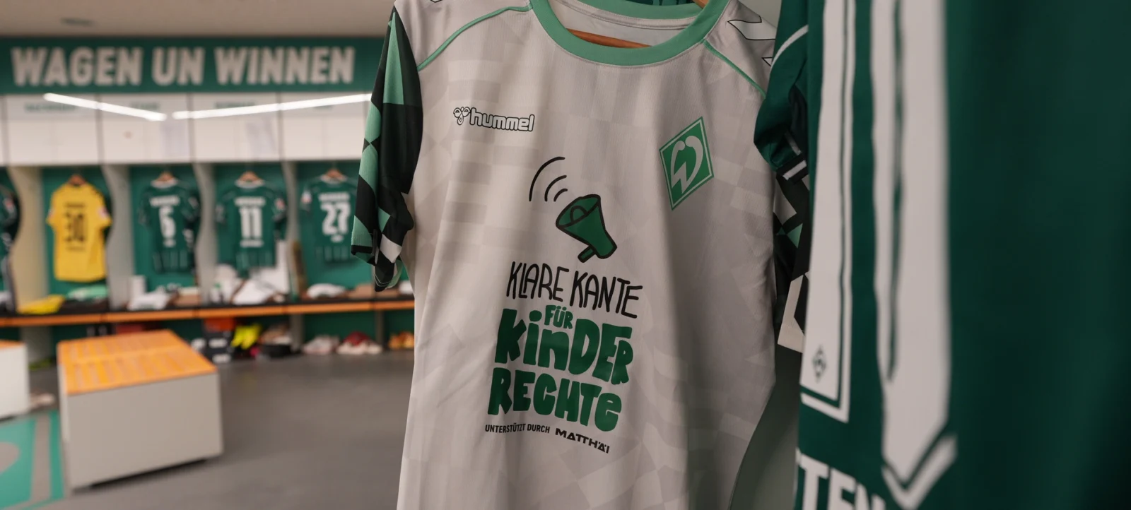 Ein Trikot mit dem Logo des Vereins Werder Bremen hängt in der Umkleidekabine. Das Trikot hat ein weißes und grünes Design mit einem Megafon und der Aufschrift "KLARE KANTE FÜR KINDERRECHTE" sowie dem Vereinslogo und dem Herstellerlogo "Hummel". Im Hintergrund sind weitere Trikots und Umkleidekabinen zu sehen.