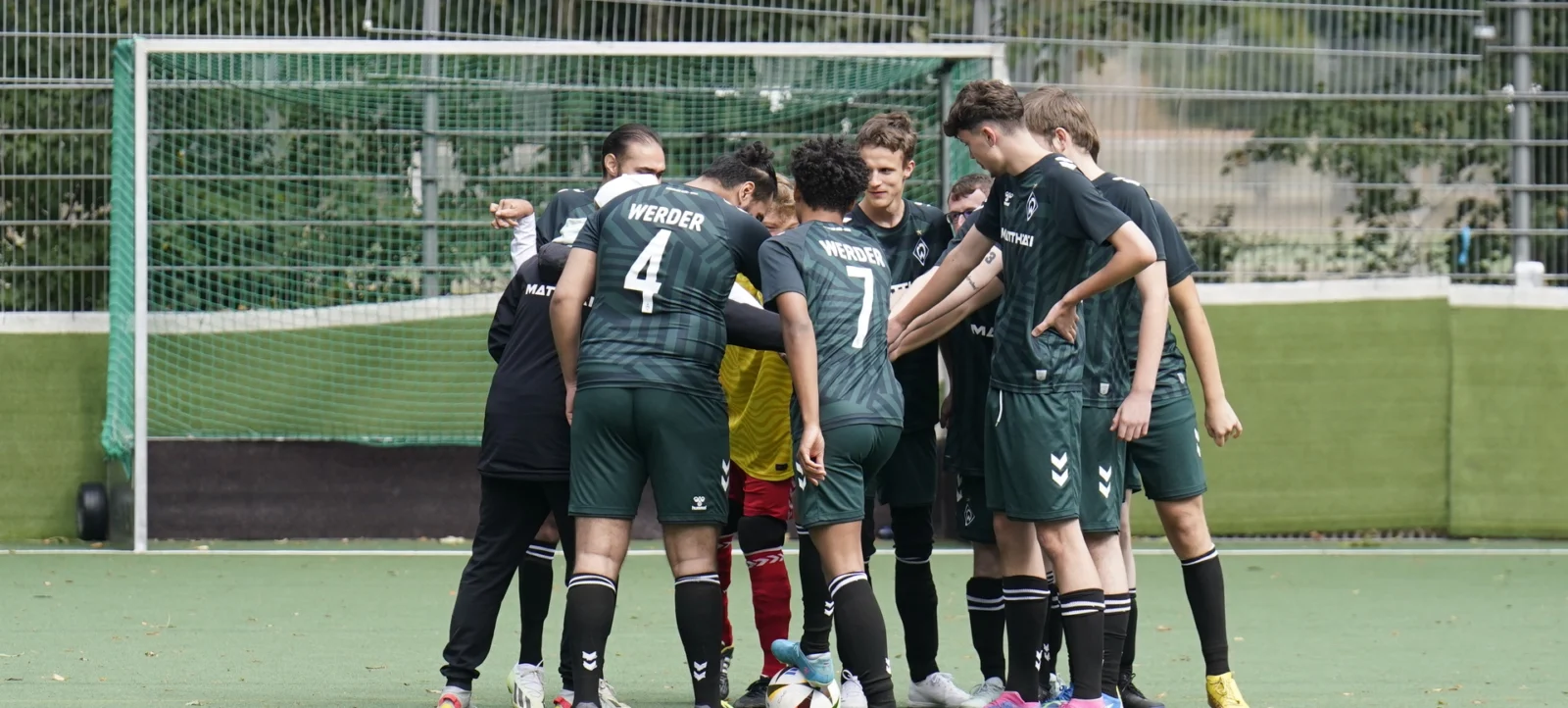 Eine Gruppe von Fußballspielern in grün-schwarzen Trikots steht im Kreis zusammen und berührt sich gegenseitig mit den Fäusten. Sie scheinen sich auf dem Spielfeld vor einem Tor zu befinden, das von einem Netz umgeben ist. Die Spieler wirken konzentriert und motiviert, möglicherweise für eine Besprechung oder eine Motivationsübung vor dem Spiel.