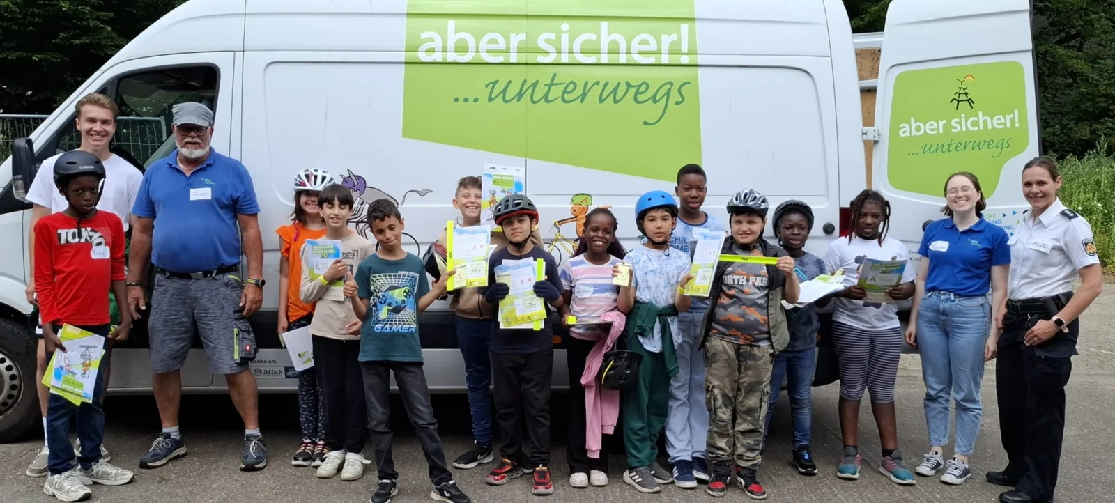 Gruppe von Kindern und Erwachsenen vor einem weißen Lieferwagen mit der Aufschrift "aber sicher! ...unterwegs". Die Kinder halten Informationsbroschüren in den Händen und lächeln in die Kamera. Die Erwachsenen tragen Uniformen und Namensschilder, was auf eine offizielle Veranstaltung oder ein Sicherheitsprogramm hinweist. Die Szene findet im Freien statt, mit Bäumen im Hintergrund.