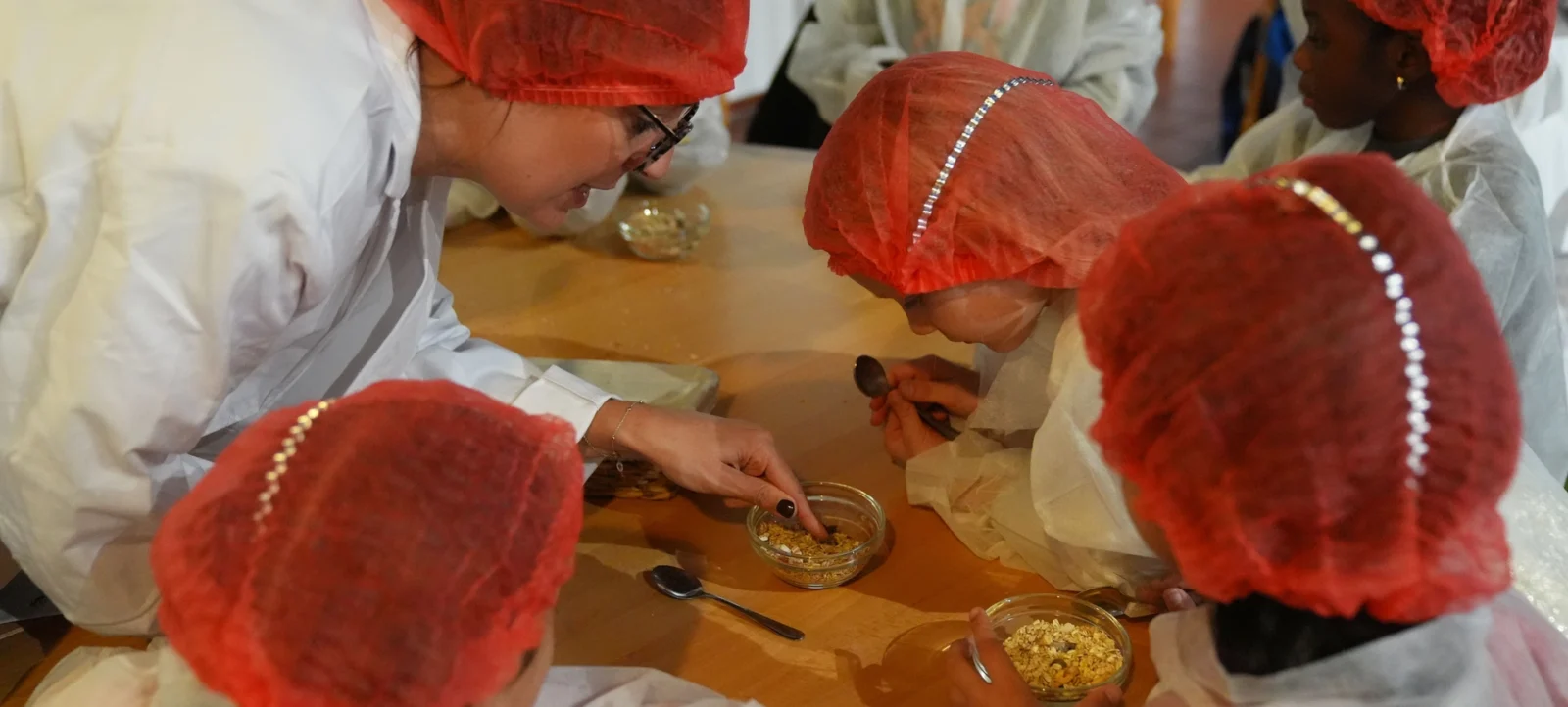 Mehrere Personen in weißen Schürzen und roten Hauben sitzen an einem Tisch und sind in eine Tätigkeit vertieft. Sie arbeiten mit kleinen Schüsseln und Löffeln, möglicherweise bei der Herstellung oder Verarbeitung von Lebensmitteln. Im Hintergrund sind weitere Personen zu sehen, die ebenfalls in die Arbeit involviert sind.