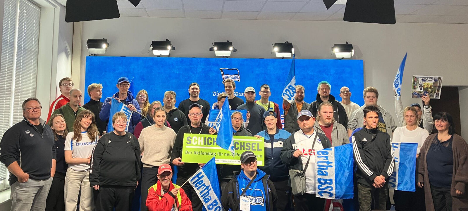 Eine Gruppe von Menschen steht vor einem blauen Hintergrund mit dem Logo von Hertha BSC. Sie halten Schilder und Fahnen, auf denen "Schichtwechsel" und "Hertha BSC" zu lesen ist. Die Personen lächeln und posieren für ein Gruppenfoto, offenbar bei einer Veranstaltung oder einem Aktionstag.