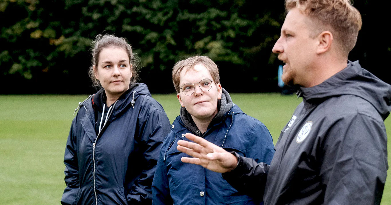 Drei Personen stehen auf einem Rasenplatz und unterhalten sich. Zwei Frauen und ein Mann sind in der Bildmitte zu sehen. Die Person rechts gestikuliert mit den Händen, während die anderen beiden aufmerksam zuhören. Alle tragen dunkle Jacken und sind vor einem Hintergrund aus Bäumen und grünem Rasen positioniert.