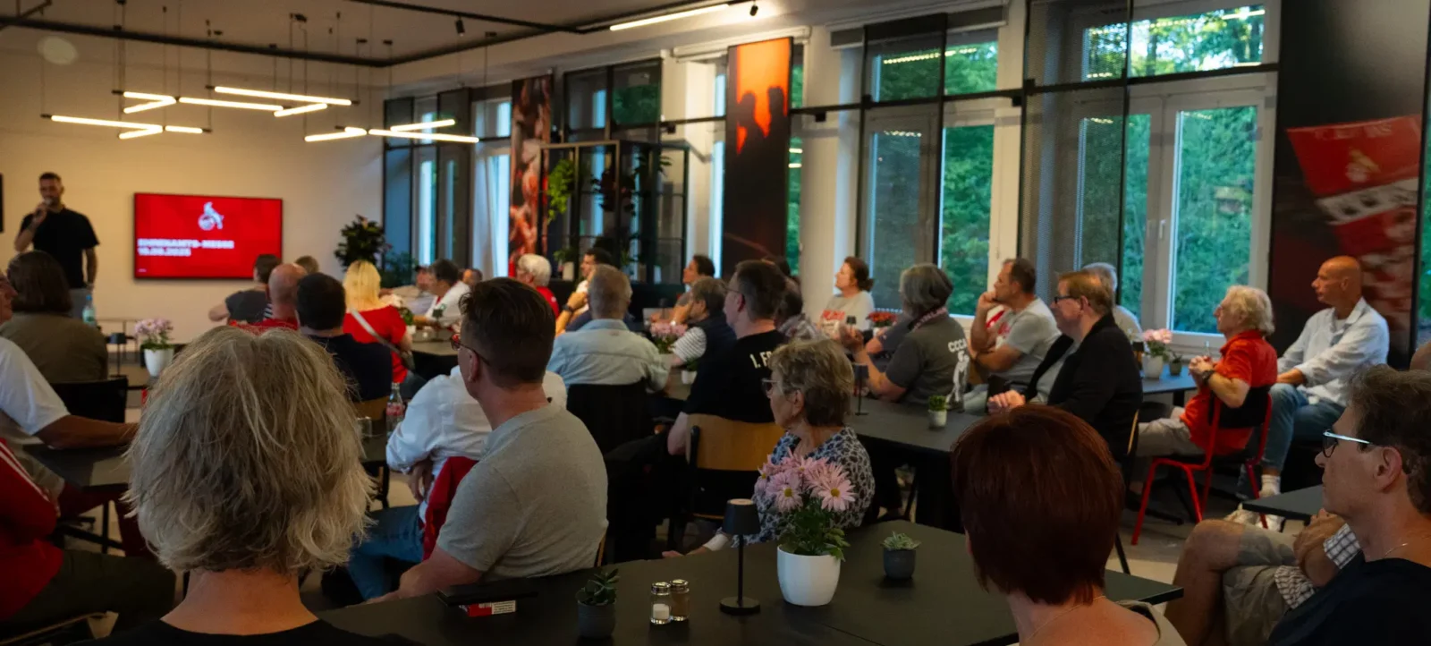 Eine Gruppe von Menschen sitzt in einem hellen Raum mit großen Fenstern und hört aufmerksam einer Person zu, die an der Vorderseite des Raumes steht. Die Wände sind mit großen Bildern von Fußballspielern dekoriert. Die Tische sind mit kleinen Pflanzen und Blumen geschmückt.