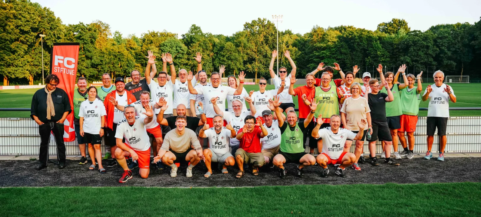 Eine Gruppe von Menschen, die für ein Gruppenfoto auf einem Fußballfeld posieren. Sie tragen T-Shirts mit dem Aufdruck "FC STIFTUNG" und andere farbenfrohe Kleidung. Die meisten von ihnen winken oder zeigen andere freudige Gesten. Im Hintergrund ist eine rote Fahne mit dem Aufdruck "FC STIFTUNG" zu sehen, sowie ein Fußballfeld und Bäume.