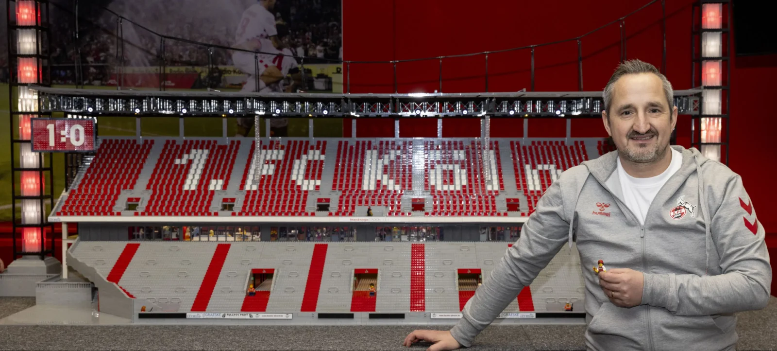 Ein Mann steht vor einem detailreichen Modell eines Fußballstadions, das aus LEGO-Steinen gebaut wurde. Das Modell zeigt das Stadion mit Sitzplätzen, einem Spielfeld und einer Anzeigetafel, die ein Spiel mit einem Stand von 1:0 anzeigt. Der Mann trägt eine graue Jacke mit einem Logo auf der Brust und lächelt in die Kamera.