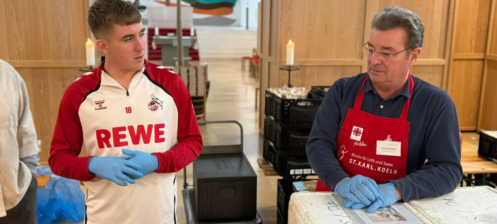 Zwei Männer in einer Küche, einer im Sportdress mit der Aufschrift "REWE" und blauem Handschuh, der andere mit einer Schürze mit dem Logo "Kirche für Leib und Seele St. Karl Köln" und blauem Handschuh, die gemeinsam an einem Projekt arbeiten.