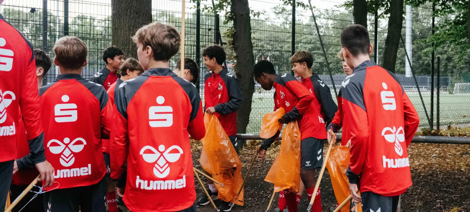 Eine Gruppe von Jugendlichen in roten und schwarzen Sportjacken mit dem Logo von Hummel steht im Freien und hält orange Müllsäcke sowie Rechen. Sie scheinen an einer Umweltaktion teilzunehmen, möglicherweise beim Aufräumen eines Bereichs.
