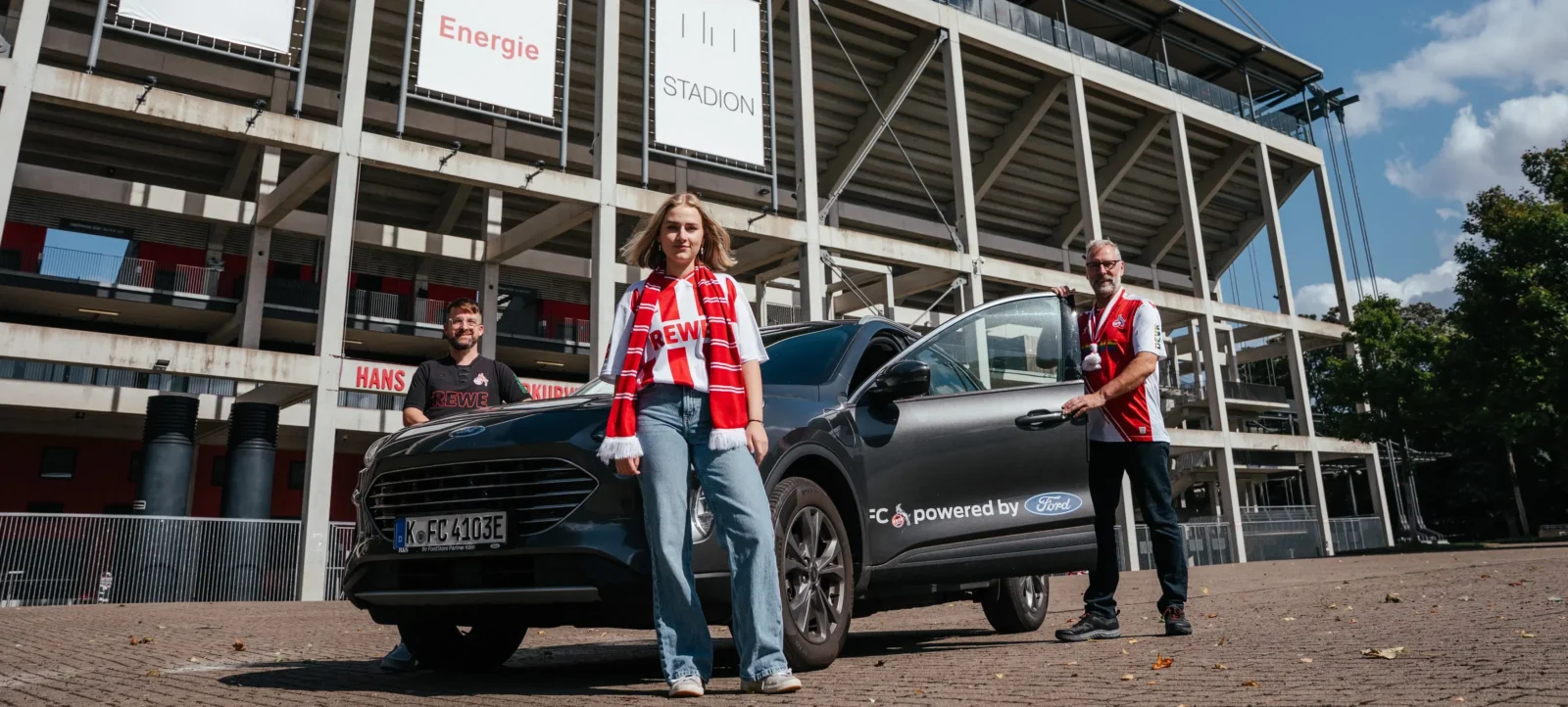 Drei Personen stehen vor einem Stadion und posieren mit einem schwarzen Ford-Kombi. Die Personen tragen Schals und T-Shirts mit dem Aufdruck "REWE". Das Auto hat ein Schild mit der Aufschrift "FC powered by Ford" an der Seite.