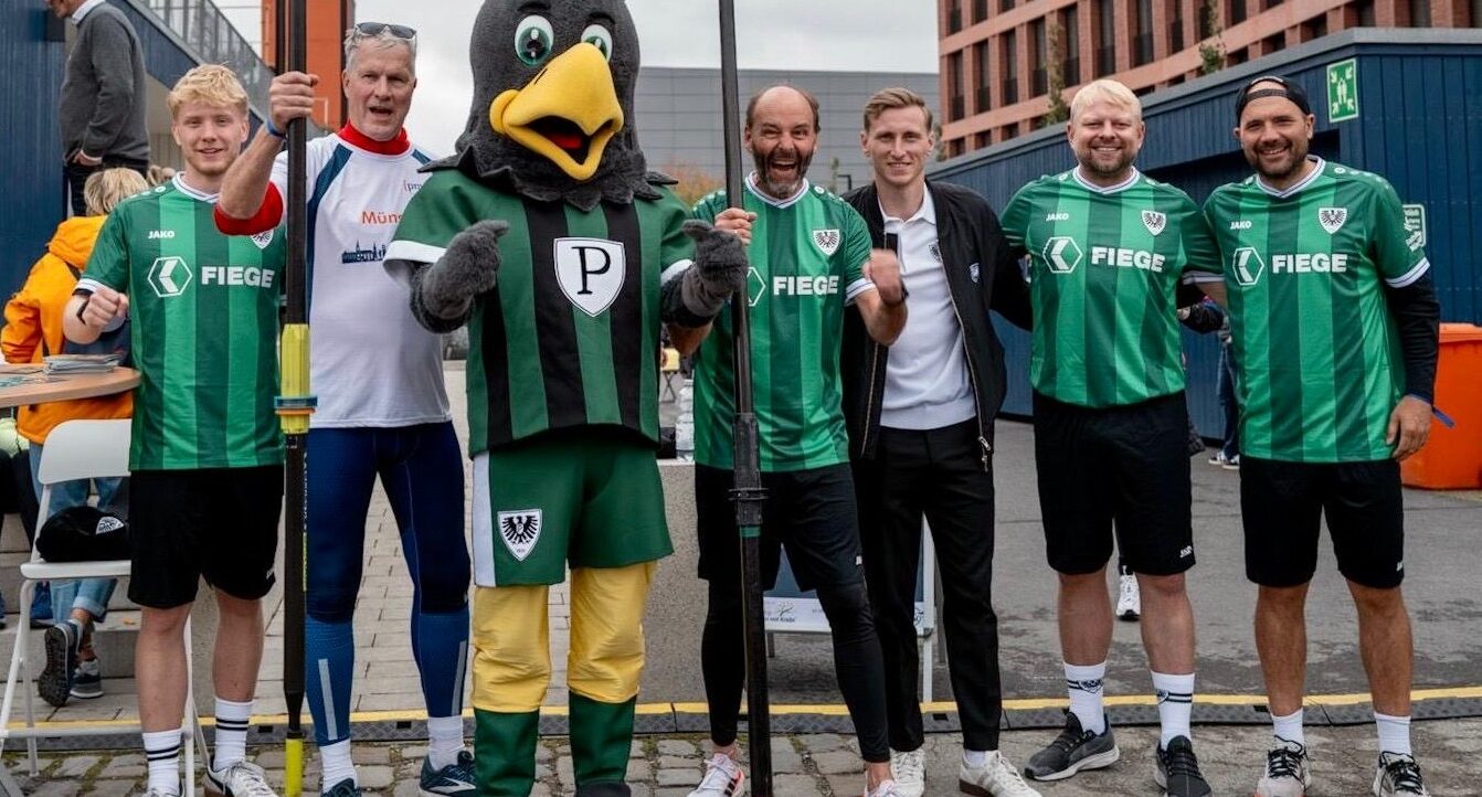 Eine Gruppe von Menschen und ein Maskottchen in Vogelkostüm stehen auf einem Steg und halten gemeinsam einen Mast. Sie tragen grüne und weiße Sportshirts mit dem Logo "Fiege" und "JAKO". Im Hintergrund sind Gebäude und eine Person in orangefarbener Kleidung zu sehen.