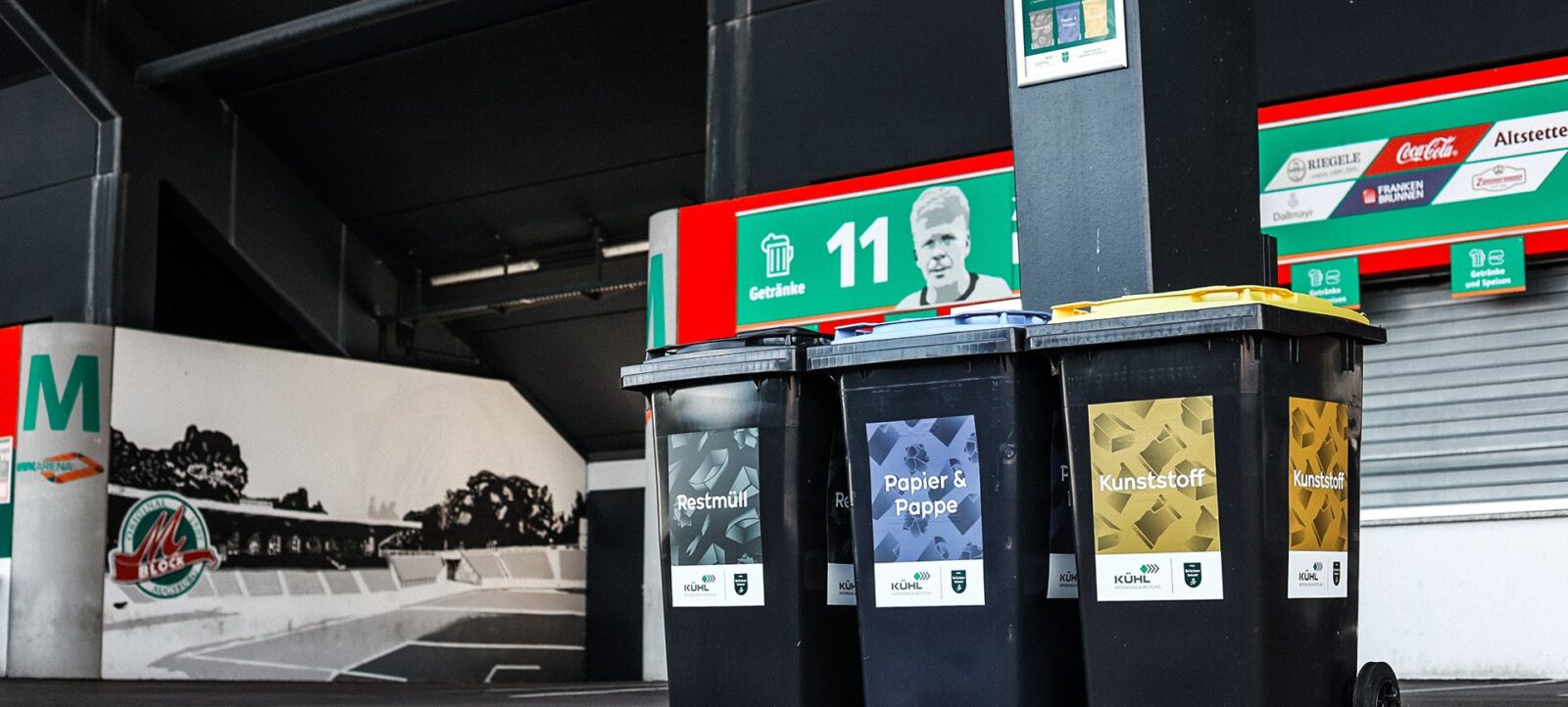 Drei Mülltonnen in einem Stadion, beschriftet mit "Restmüll", "Papier & Pappe" und "Kunststoff" für die Mülltrennung.