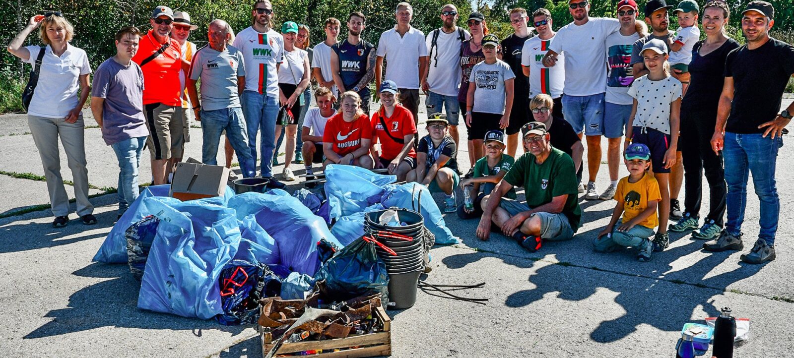 Eine Gruppe von Menschen, darunter Erwachsene und Kinder, steht für ein Gruppenfoto auf einem Parkplatz. Sie tragen verschiedene T-Shirts, einige mit Aufdrucken wie "WWK" und "AUGSBURG". Vor der Gruppe liegt eine Sammlung von Müllsäcken und gesammelten Abfällen auf dem Boden, darunter blaue und schwarze Säcke, ein Eimer und verschiedene andere Gegenstände. Die Gruppe scheint an einer Müllsammelaktion teilgenommen zu haben, möglicherweise im Rahmen eines Nachhaltigkeitsprojekts.
