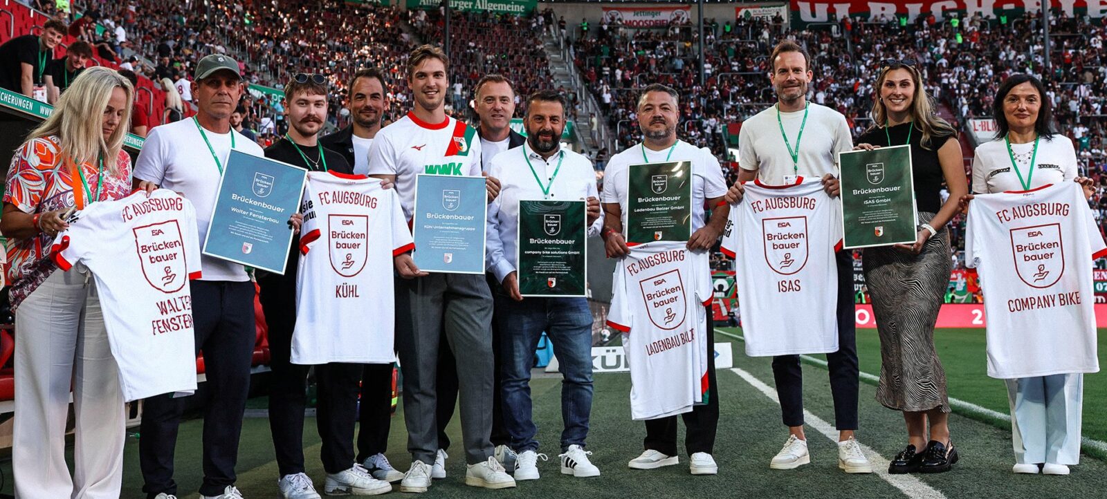 Eine Gruppe von Personen steht auf dem Spielfeld eines Fußballstadions und hält Trikots und Urkunden des FC Augsburg. Die Trikots tragen den Schriftzug "Brückenbauer" und die Namen verschiedener Unternehmen. Im Hintergrund sind zahlreiche Zuschauer auf den Rängen zu sehen.