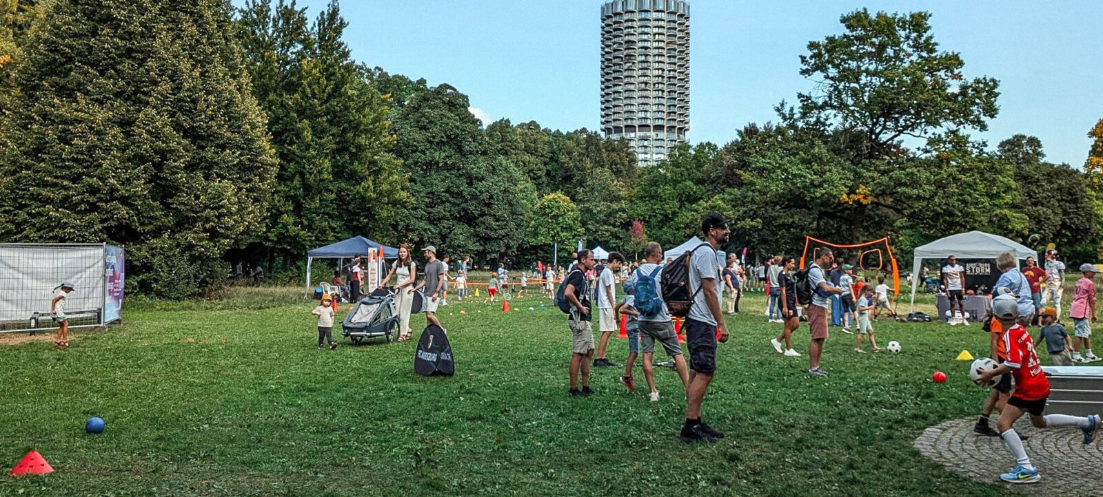 Ein belebter Park mit vielen Menschen, die an verschiedenen Aktivitäten teilnehmen. Im Vordergrund sind Kinder in roten Trikots zu sehen, die mit Bällen spielen. Erwachsene und Kinder sind auf dem Rasen verteilt und nehmen an verschiedenen Stationen teil, darunter ein Zelt und ein Tor. Im Hintergrund steht ein großes, markantes Gebäude. Der Park ist von vielen Bäumen umgeben, und das Wetter scheint klar und sonnig zu sein.