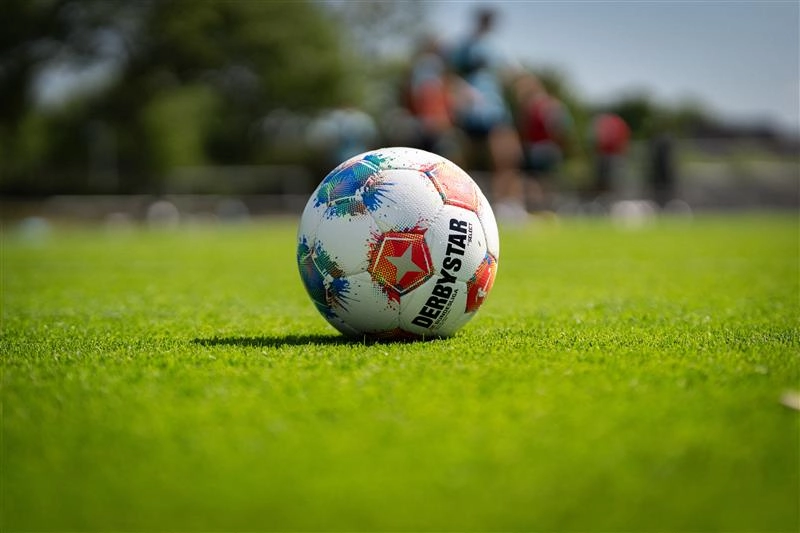 Ein Derby Star Fußball liegt auf einem grünen Rasenplatz. Im Hintergrund sind unscharf Menschen zu erkennen.