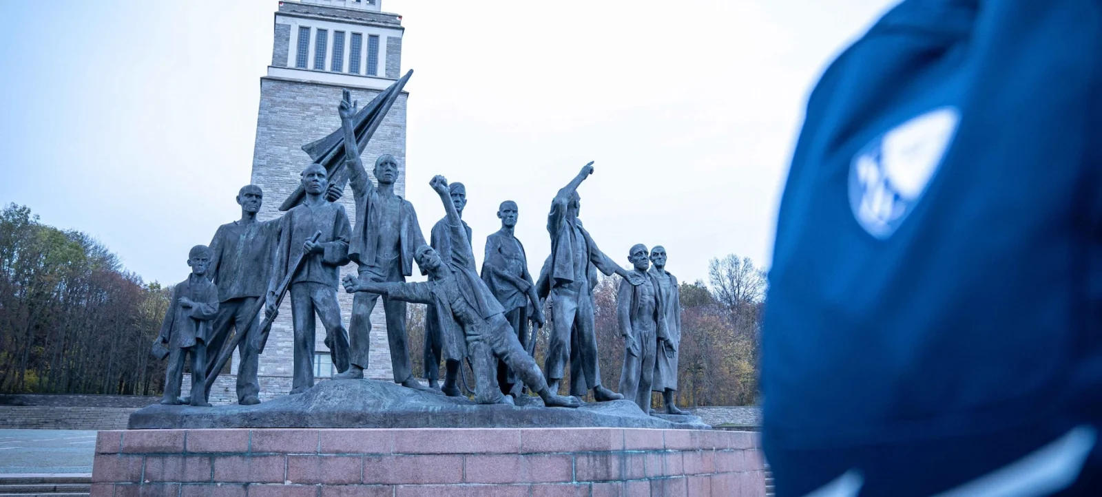 Eine Gruppe von Menschen in einer Skulptur vor einem hohen Turm mit der Aufschrift MCMLXV. Die Skulptur zeigt verschiedene Figuren in dynamischen Posen, einige halten Fahnen und andere gestikulieren. Im Vordergrund ist ein unscharfes blaues Trikot mit einem weißen Emblem zu sehen.
