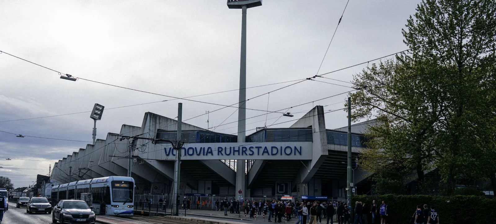 Außenansicht des Vonovia Ruhrstadions mit einem hohen Flutlichtmast im Vordergrund. Ein Straßenbahnwagen fährt auf der Straße vorbei, und zahlreiche Menschen sind auf dem Gehweg zu sehen. Der Himmel ist bewölkt, und Bäume rahmen die Szene ein.