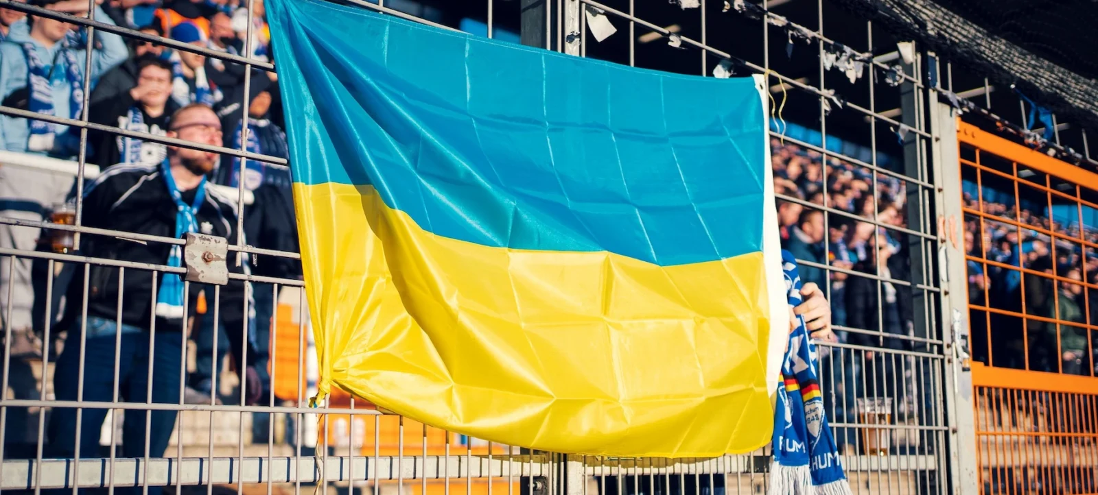 Ein großes blau-gelbes Flagge, die an einem Gitter befestigt ist, wird von einem Fan im Stadion gehalten. Im Hintergrund sind weitere Fans zu sehen, die das Spiel verfolgen.