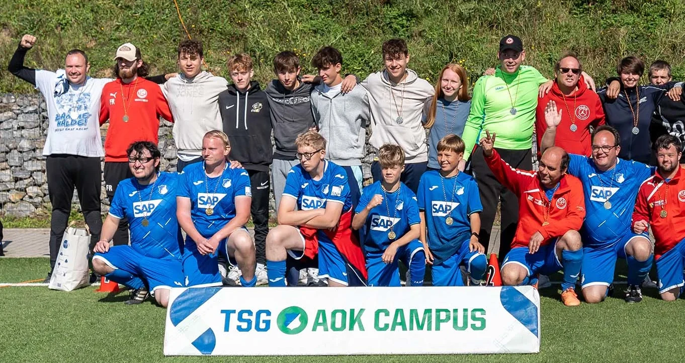Eine Gruppe von Menschen, darunter Kinder und Erwachsene, posieren für ein Gruppenfoto auf einem Rasenfeld. Die Kinder in der vorderen Reihe tragen blaue T-Shirts mit dem Aufdruck "SAP" und Medaillen um den Hals. Die Erwachsenen und einige der Kinder in der hinteren Reihe tragen verschiedene farbige Kleidung und einige von ihnen haben ebenfalls Medaillen um den Hals. Vor der Gruppe liegt ein Banner mit der Aufschrift "TSG AOK CAMPUS". Die Atmosphäre wirkt fröhlich und gesellig.
