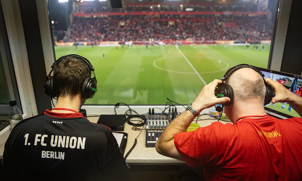 Zwei Personen in der Pressetribüne eines Fußballstadions, die ein Spiel des 1. FC Union Berlin verfolgen. Beide tragen Kopfhörer und sind auf das Geschehen auf dem Spielfeld konzentriert. Eine Person trägt ein schwarzes T-Shirt mit der Aufschrift "1. FC Union Berlin", die andere ein rotes T-Shirt mit der Aufschrift "Europa League". Im Vordergrund sind technische Geräte wie ein Mischpult und ein Bildschirm zu sehen.