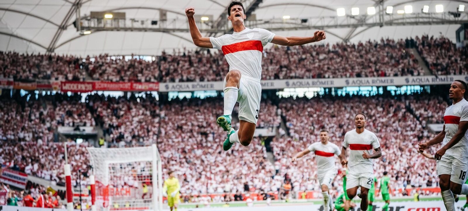 Ein Fußballspieler in weißem Trikot mit rotem Streifen springt freudig in der Luft, nachdem er ein Tor geschossen hat. Im Hintergrund sind seine jubelnden Mitspieler und eine große Menge begeisterter Zuschauer im Stadion zu sehen.