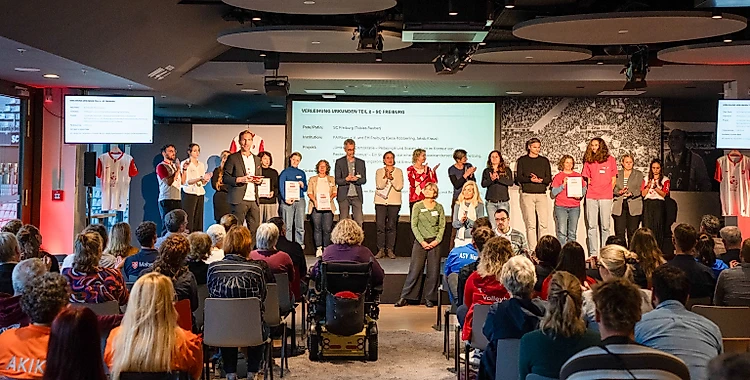 Eine Gruppe von Menschen steht auf einer Bühne und hält Urkunden oder Zertifikate in den Händen. Sie scheinen bei einer Veranstaltung oder einem Workshop zu sein, möglicherweise im Zusammenhang mit Nachhaltigkeit oder Umweltinitiativen. Die Bühne ist mit einem großen Bildschirm im Hintergrund ausgestattet, auf dem der Text "Sustainability Champions" und weitere Informationen zu sehen sind. Das Publikum sitzt vor der Bühne und beobachtet die Veranstaltung. Die Atmosphäre wirkt feierlich und motivierend.