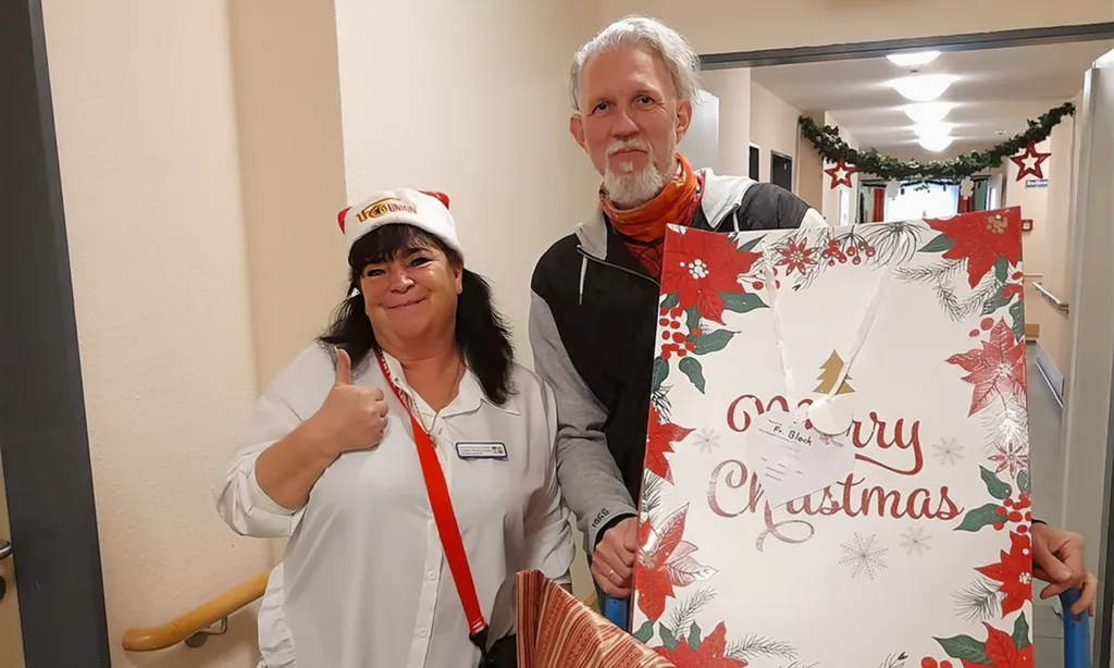 Zwei Personen in einem Flur, die ein Weihnachtskarten-Plakat mit der Aufschrift "Merry Christmas" halten. Eine Person trägt eine Weihnachtsmann-Mütze und zeigt mit einem Daumen nach oben, während die andere Person lächelt. Der Flur ist mit Weihnachtsdekorationen geschmückt.