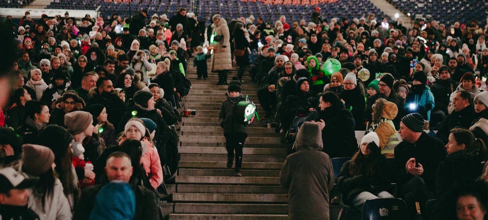 Eine große Menschenmenge nimmt an einem Laternenumzug in einem Stadion teil. Die Zuschauer sind auf den Sitzplätzen und Treppen verteilt und tragen warme Kleidung, darunter Mützen und Jacken. Einige Personen halten Laternen, die grün leuchten. Die Atmosphäre wirkt festlich und gemütlich.