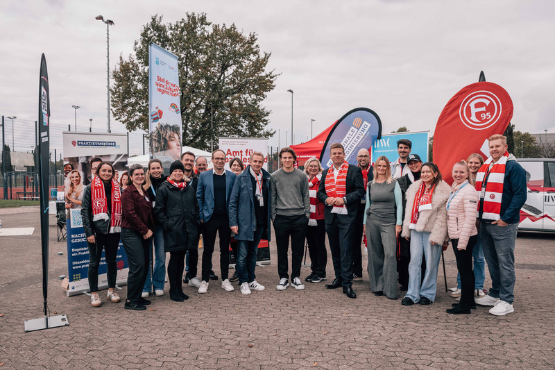 Gruppenfoto von Menschen vor einem Stadion. Die Gruppe steht auf einem gepflasterten Bereich vor mehreren Fahnen und Bannern, darunter eines mit dem Logo des 1. FC Köln. Die Personen tragen verschiedene Kleidungsstücke, einige mit Schals in den Vereinsfarben. Im Hintergrund sind Bäume und ein Teil des Stadions zu sehen.