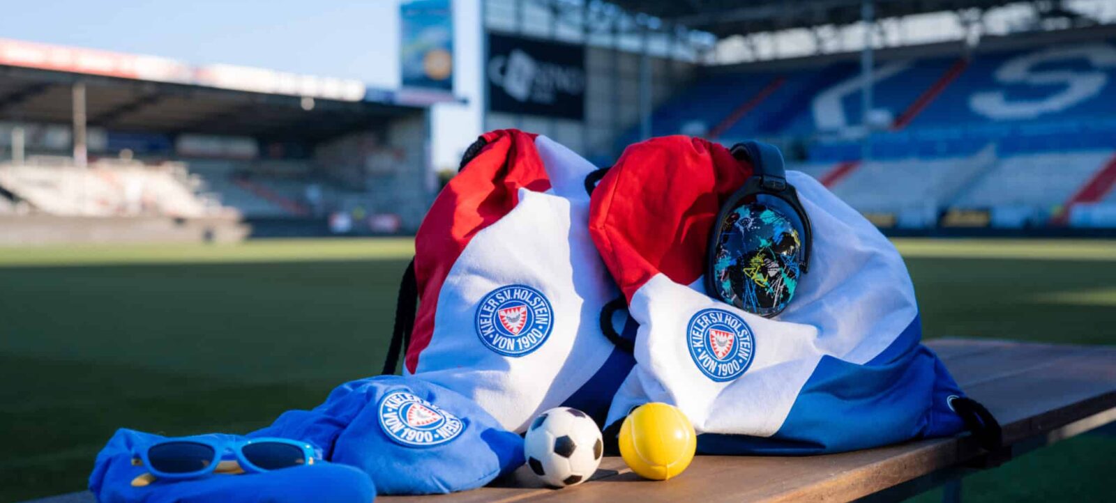 Auf einer Bank vor einem Fußballstadion liegen verschiedene Gegenstände: eine rote und weiße Tasche mit dem Logo des Vereins "Wester St. Tönnis von 1900", eine blaue Mütze, eine schwarze Sonnenbrille, ein weißer und schwarzer Fußball und eine gelbe Tennisball.