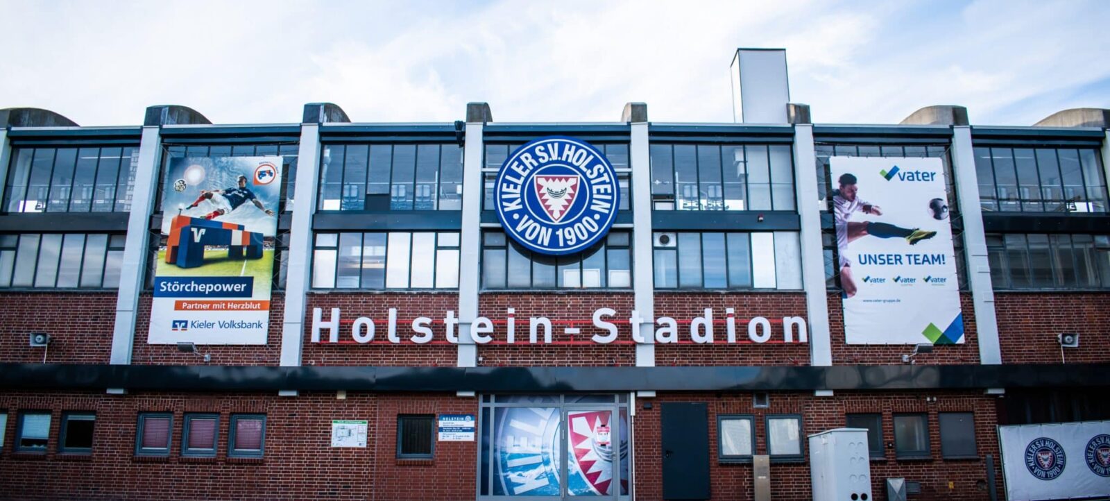 Fassade des Holstein-Stadions mit dem Vereinslogo von Kieler SV Holstein von 1900. Zwei Werbebanner sind zu sehen: eines mit dem Slogan 'Störchepower' und dem Logo der Kieler Volksbank, das andere mit dem Slogan 'Unser Team!' und dem Logo der vater Gruppe. Der Himmel ist leicht bewölkt.