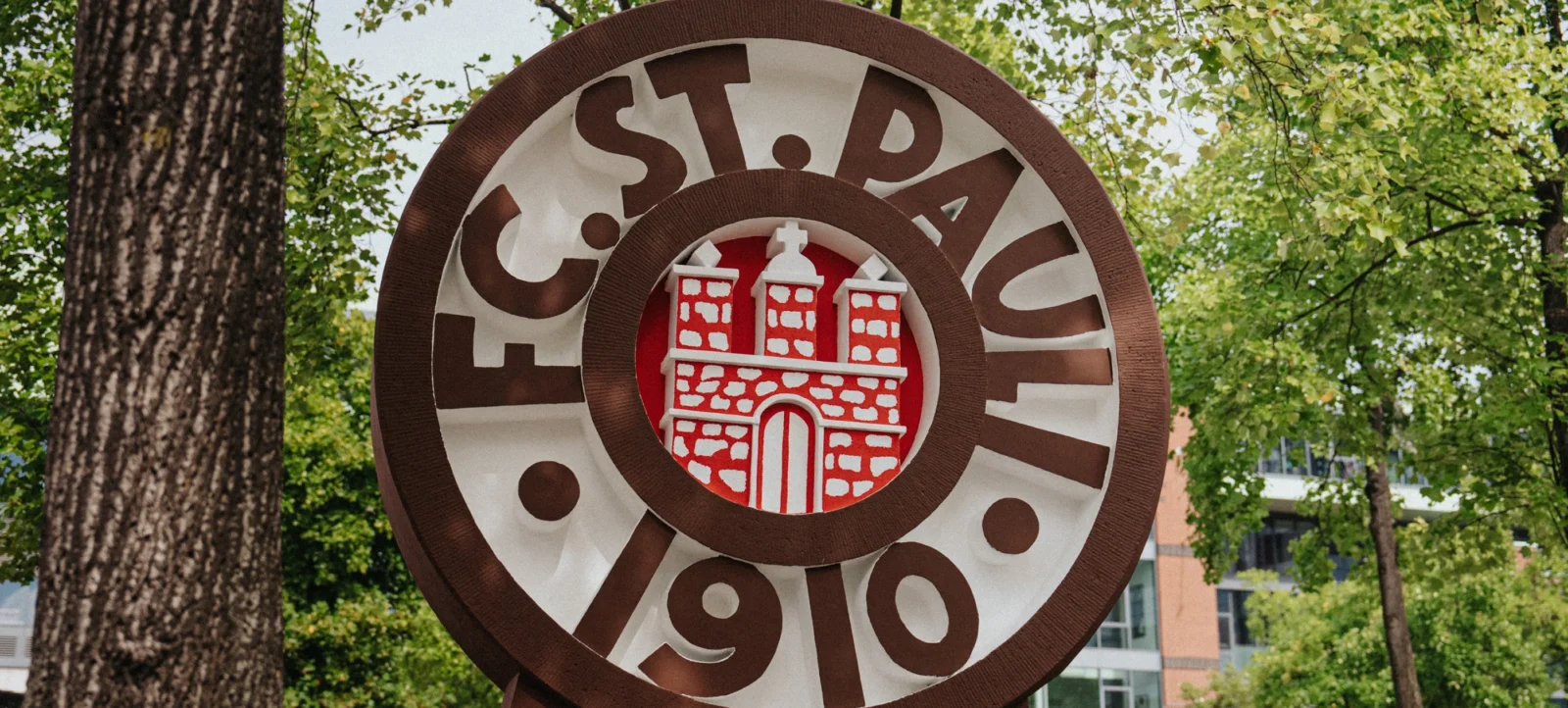 Ein runder, dreidimensionaler Schild mit dem Wappen des FC St. Pauli. Das Wappen zeigt ein rotes Gebäude mit drei Türmen und einem Tor. Um das Wappen herum steht der Schriftzug "FC ST. PAULI 1910" in braunen Buchstaben. Das Schild ist an einem Baum in einem grünen, bewaldeten Gebiet befestigt.