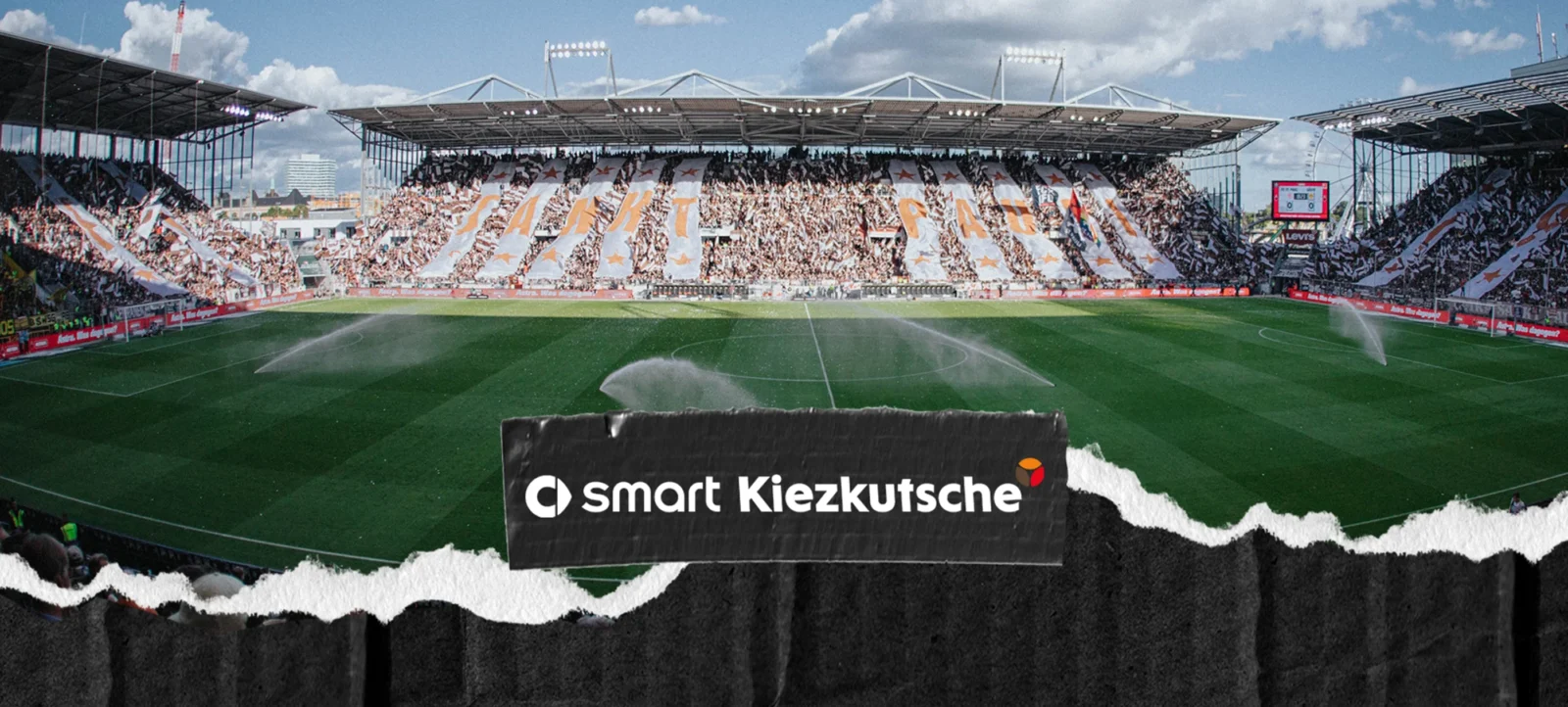 Ein Fußballstadion mit einer großen Zuschauermenge, die auf den Rängen sitzt. Die Fans halten Schilder hoch, auf denen "WIR HABEN DICH" zu lesen ist. Im Vordergrund befindet sich ein Werbebanner mit der Aufschrift "smart Kiezkutsche". Das Spielfeld ist grün und wird von Sprinklern bewässert.