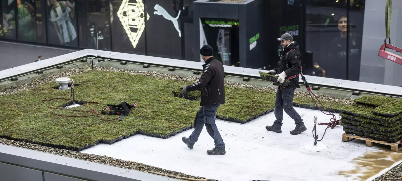 Zwei Arbeiter in schwarzer Kleidung und Mützen stehen auf einem grünen Dach und führen Wartungsarbeiten durch. Sie sind mit Handschuhen und Werkzeugen ausgestattet. Im Hintergrund ist ein Gebäude mit einem großen Logo und einem Puma-Logo zu sehen.