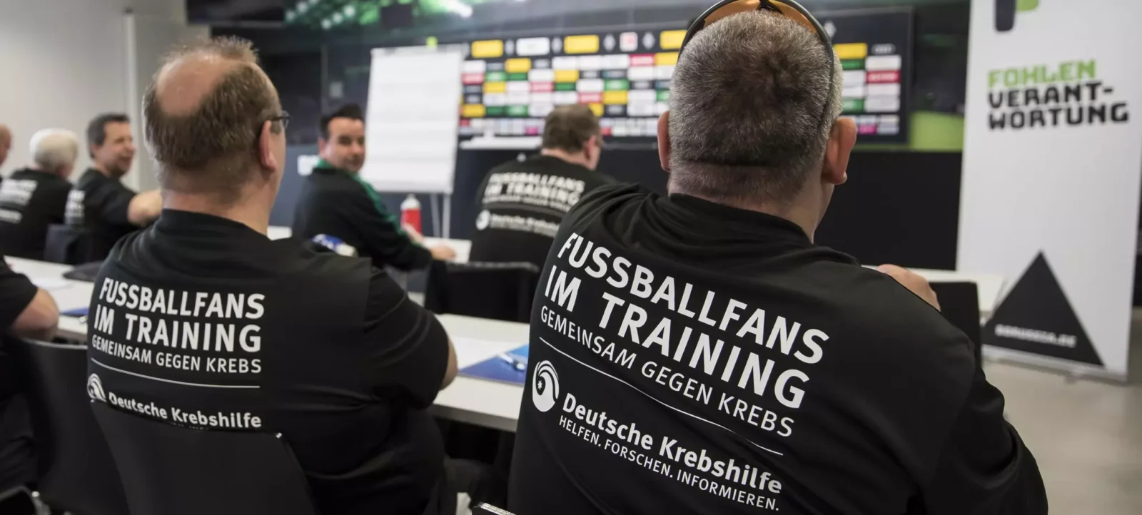 Zwei Männer mit schwarzen T-Shirts, auf denen 'Fußballfans im Training gemeinsam gegen Krebs' steht, sitzen in einem Raum und schauen auf eine große Anzeigetafel. Im Hintergrund sind weitere Personen zu sehen, die ebenfalls an der Veranstaltung teilnehmen. Die Veranstaltung scheint sich auf Krebsprävention oder -bewusstsein zu konzentrieren und wird von der Deutschen Krebshilfe unterstützt.