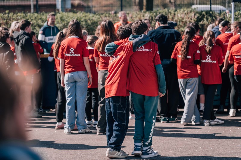 Eine Gruppe von Menschen in roten T-Shirts mit dem Aufdruck "TARGOBANK" steht im Freien und scheint sich auf eine Aktivität oder ein Event vorzubereiten. Die Personen sind in einer lockeren Gruppe angeordnet und unterhalten sich miteinander. Im Hintergrund sind weitere Menschen und ein Zaun zu sehen.