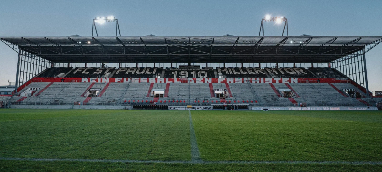 Ein Fußballstadion bei Dämmerung. Die Tribüne zeigt eine große Schrift mit den Worten "HEIMRUHE GIBT'S NUR IN EINER STADT". Das Spielfeld ist grün und gut beleuchtet, während die Umgebung langsam dunkel wird.