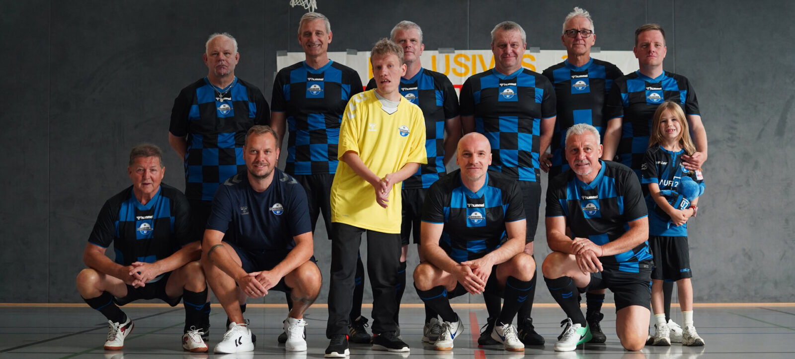 Eine Gruppe von Menschen, darunter Erwachsene und ein Kind, posieren für ein Gruppenfoto in einer Sporthalle. Die meisten tragen blaue und schwarze Sportshirts mit einem Logo, während eine Person ein gelbes T-Shirt trägt. Sie sind vor einem Banner mit der Aufschrift "USIV" aufgestellt. Die Atmosphäre wirkt fröhlich und gesellig, was auf ein inklusives Sportereignis hindeutet.