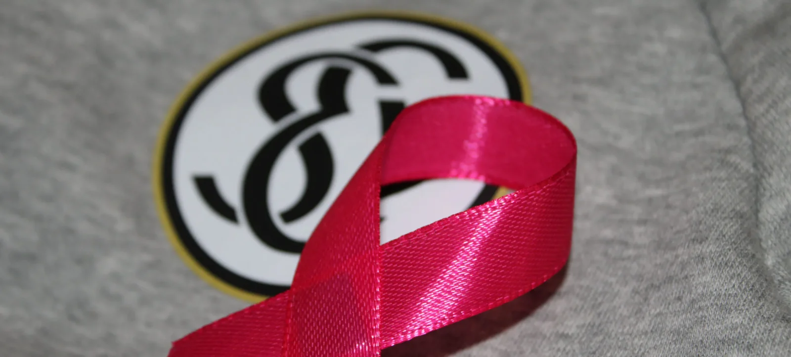 Ein graues T-Shirt mit einem runden Aufnäher, der das Logo "FC" in schwarzer Schrift auf weißem Hintergrund zeigt. Ein rotes Band ist über dem Aufnäher befestigt und symbolisiert Unterstützung für eine soziale oder gesundheitliche Initiative.