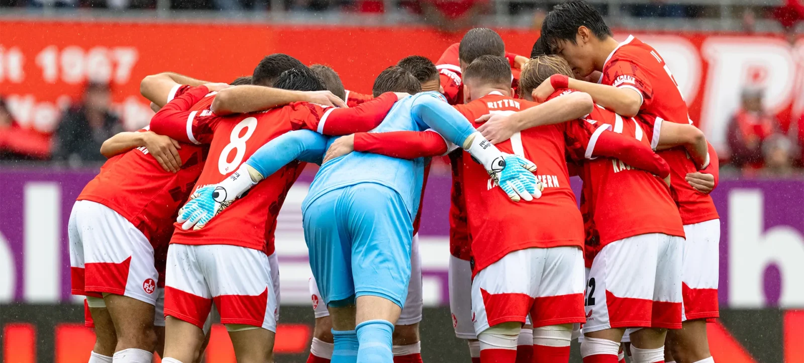 Eine Gruppe von Fußballspielern in roten Trikots und einem Torwart in blauem Trikot umarmen sich auf dem Spielfeld, vermutlich nach einem Tor oder einem wichtigen Spielmoment. Der Hintergrund zeigt eine Stadionatmosphäre mit teilweise erkennbaren Zuschauern und Werbebanden.