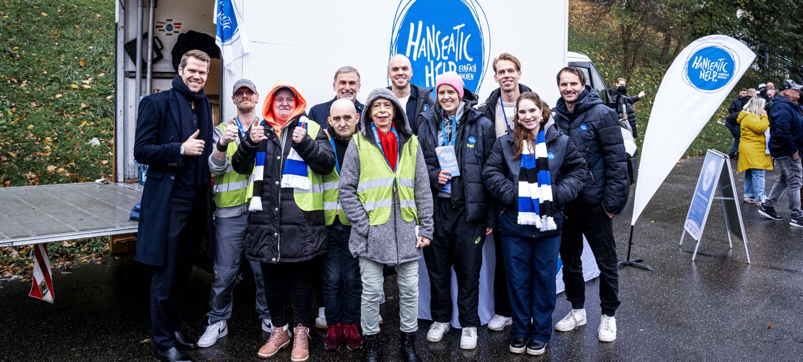 Eine Gruppe von Menschen, darunter Freiwillige in gelben Westen und Mitglieder des HSV, stehen vor einem mobilen Stand mit dem Logo "Hanseatic Help". Sie lächeln und machen Daumen-hoch-Gesten, während sie für ein Gruppenfoto posieren. Der Hintergrund zeigt einen bewaldeten Bereich und weitere Personen im Hintergrund.