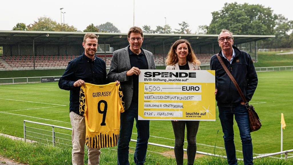 Vier Personen stehen auf einem Fußballfeld und halten ein Scheck über 4.500 Euro für die BVB-Stiftung "leuchte auf". Eine Person hält zusätzlich ein gelb-schwarzes Trikot mit der Nummer 9 und dem Namen "GUIRASSY". Die Szene spielt auf einem Rasenplatz mit Tribünen im Hintergrund.