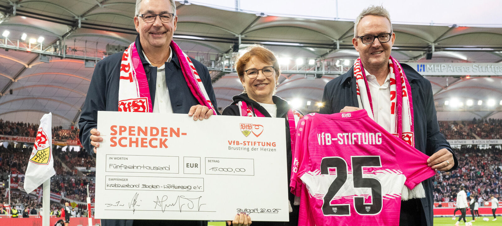 Drei Personen stehen auf einem Fußballfeld und halten einen großen Spendenscheck der VfB-Stiftung im Wert von 15.000 Euro für den Krebsverband Boden-Württemberg e.V. Sie tragen schwarze Jacken mit roten und weißen Schal-Schals und lächeln in die Kamera. Im Hintergrund ist die MHP Arena Stuttgart mit vielen Zuschauern zu sehen.